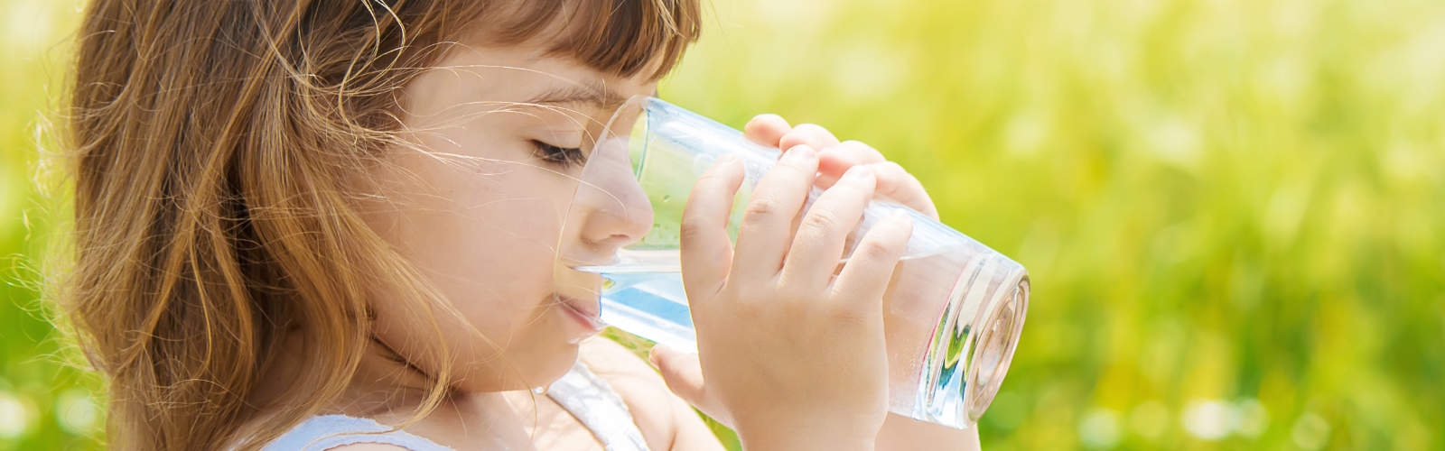 Girl drinking glass of water outdoors in green field.
